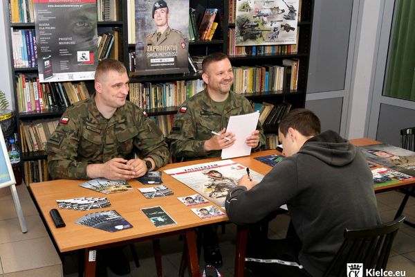 Młody mężczyzna siedzi przy stole naprzeciwko dwóch żołnierzy