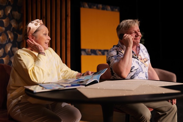 Zdjęcie przedstawia scenę  z komedii pt. „Szczęściarze”. Dwoje aktorów siedzą przy stole, kobieta czyta gazetę, a mężczyzna rozmawia przez telefon.