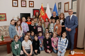 Kilkanaścioro dzieci w gabinecie prezydenta Kielc oraz cztery osoby dorosłe. W tle stoją flagi.