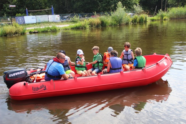 Grupa dzieci ubrana w kapoki płynie na czerwonym pontonie z mężczyzną.