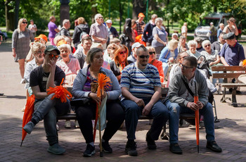 Grupa osób siedzi na łąwkach przed muszlą w Parku Miejskim. 