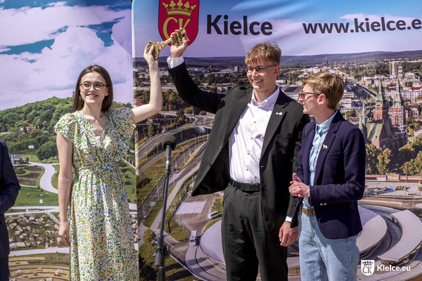 Młoda kobieta w sukience i mężczyzna w garniturze trzymają w dłoniach duży miedziany klucz i podnoszą go do góry. Obok stoi prezydentka Kielc Agata Wojda.