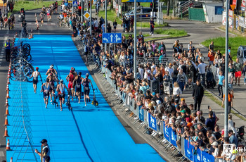 Zdjęcie z lotu ptaka, po prawej stronie kilkaset osób dopinguje zawodników, po środku grupa triathlonistów prowadzi rowery w strefie zmian.