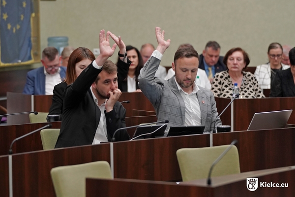 Na pierwszym planie troje radnych podnosi ręce. Na drugim planie w ławach na sali sesyjnej siedzi kilka osób.