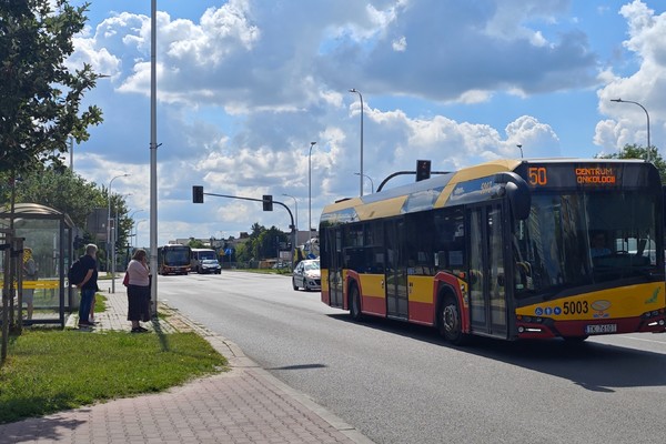 Autobus miejski jedzie ulicą, po lewej stronie przystanek, na którym stoją trzy osoby.
