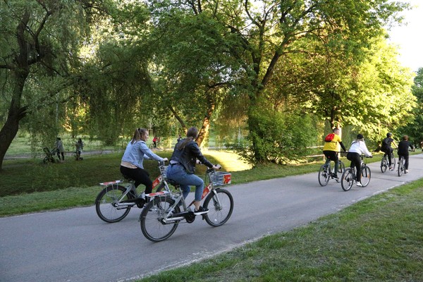 Dwie kobiety jadą na miejskich rowerach po ścieżce w Dolinie Silnicy, przed nimi jedzie czterech innych rowerzystów. Na drugim planie drzewa, spacerowicze i osoby na rowerach