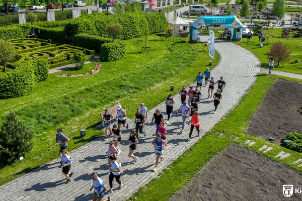 Grupa biegaczy biegnie alejką Ogrodu Botanicznego w Kielcach. Na drugim planie brama startu biegu