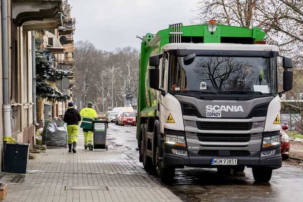 Śmieciarka stoi przy kamienicach, na chodniku dwóch mężczyzn, jeden z nich ciągnie pojemnik